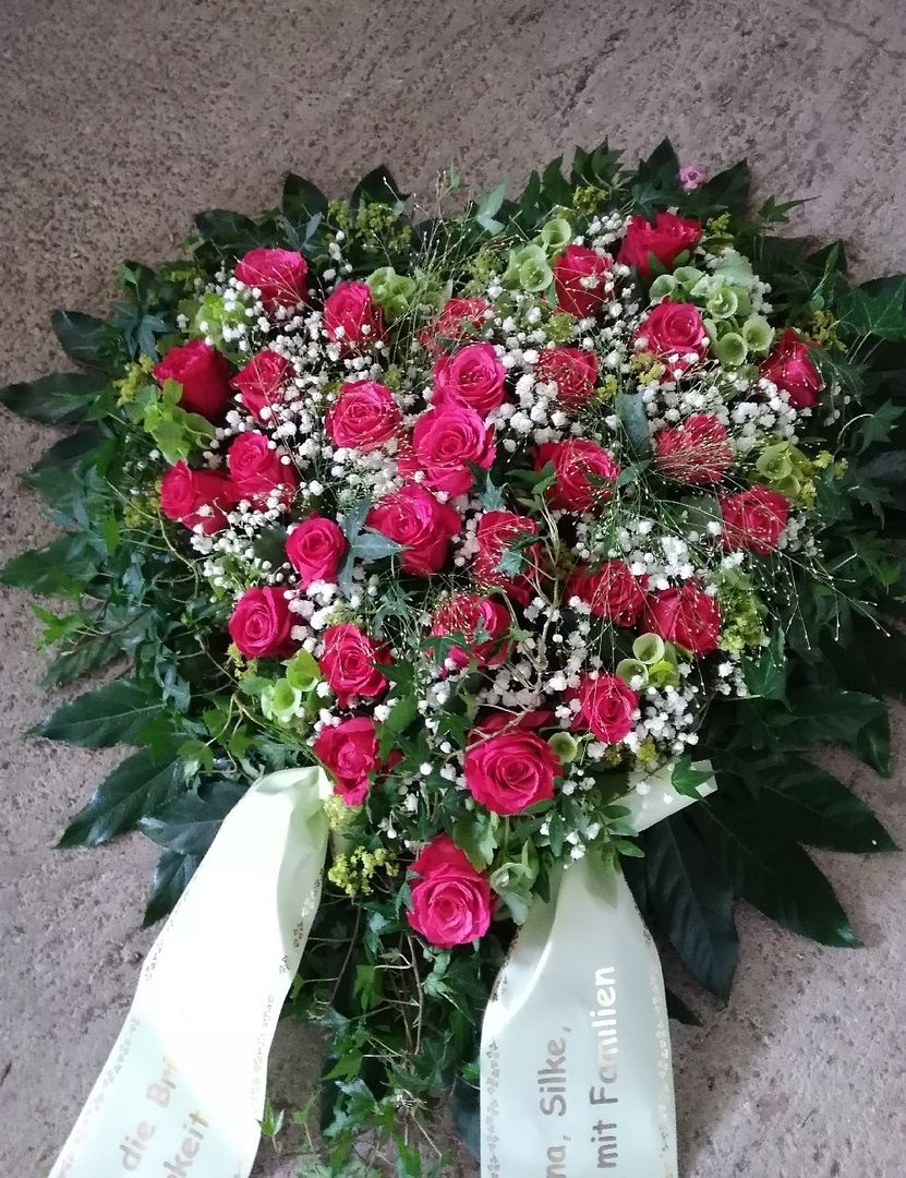 Herzförmiges Trauergesteck aus roten Rosen und weißem Schleierkraut mit grüner Umrandung und beschrifteten Schleifen